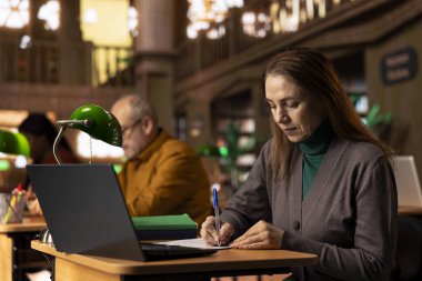 Akademik arşivlenmiş belgeler ve kitaplarla çevrili üniversite diploması için sınıf çalışması ve inceleme materyalleri hazırlayan olgun bir yetişkin. Çalışma alanında sessiz bir öğrenim ortamı.