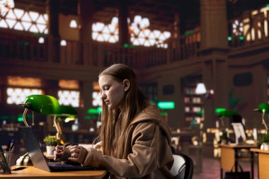 Üniversite öğrencisi, yeşil bir lambanın altında, kampüsteki üniversite kütüphanesini kullanarak önemli bir okul projesine hazırlanıyor. Genç kız bilgisayarında veri topluyor.