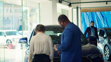 Müşteriler araba galerisine geliyor, güzel mavi modellere bakıyor, ideal yatırım seçeneklerini seçmeye çalışıyorlar. Müşteriler galeriye giriyor, araç satın almak istiyorlar, kamera A.