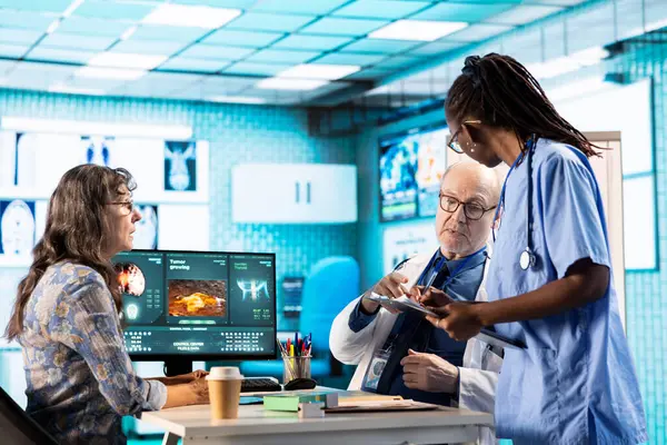 Yaşlı kadını muayene ederken kıdemli doktora yardımcı olan Afrikalı Amerikalı hemşire röntgen sonuçlarını ve tanı raporlarıyla dosyaları inceliyor. Tıbbi personel profesyonel bir yaklaşım gösteriyor..