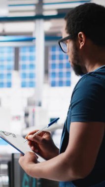 Güneş paneli fabrika incelemesinde dikey video müfettişi imalat ekipmanı ve üretim süreci, belge dosyalarını kontrol ediyor. Siyahi adam tesiste çevre dostu fotovoltaikleri sağlıyor. Kamera A