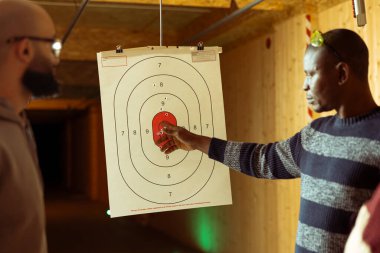 Galeri çalışanı, uygun tekniği kullanarak hedefleri nasıl vuracağını anlatıyor. Atış poligonu profesyoneli nişan alıyor, adama tabancayla nasıl ateş edileceğini öğretiyor.
