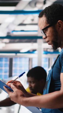 Fotovoltaik fabrikasındaki dikey video mühendisi, güneş panelleri montaj hatlarının performansını gözden geçirir, ince ayar için gerekli evrakları kontrol eder. Güneş pili makinelerini denetleyen denetçi. Kamera A.