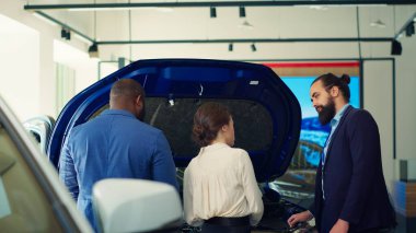 Müşterilere araç özellikleri hakkında derin bilgi sağlayan satış elemanı kaput altında kaliteli parçalar gösteriyor. Showroom temsilcisi müşterilerle arabanın içindeki motor gücü ve kamera A hakkında konuşuyor.