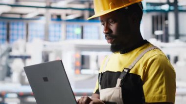 Fotovoltaik fabrikası teknisyeni dizüstü bilgisayar kullanarak üretim akışını optimize ediyor, şapka takıyor. Sürdürülebilir enerji güneş panelleri cihaz üzerindeki üretim sürecini izleyen bitki uzmanları. Kamera A.