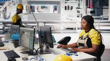 Siyah fabrika çalışanı, üretim bandı makinelerinin yanındaki masada oturan güneş sistemi performansını denetliyor. Kadın teknisyen güneş enerjisi akışını analiz eder ve sistem yazılımını günceller. Kamera B.