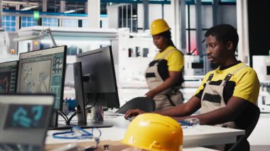 Siyah mühendis otomatikleştirilmiş fabrikada güneş yazılımı ayarları üzerinde çalışarak üretim sürecini optimize ediyor. Dijital araçlar ve endüstriyel robotlar kullanılarak optimal sistem yapılandırmasını sağlar. Kamera B.