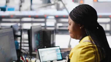 Siyah fabrika çalışanı, üretim bandı makinelerinin yanındaki masada oturan güneş sistemi performansını denetliyor. Kadın teknisyen güneş enerjisi akışını analiz eder ve sistem yazılımını günceller. Kamera A.