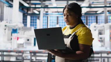 Fotovoltaik fabrikasındaki kadın mühendis arızalı sistemleri tespit etmek için dizüstü bilgisayarda izleme yazılımı kullanıyor. Güneş panelleri fabrika işçisi üretiyor. Çalışma saatlerini azaltıyor. Kamera A.