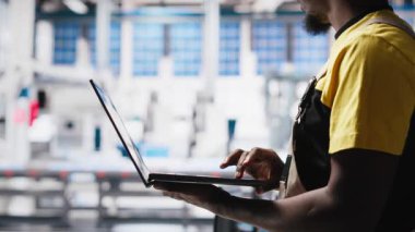 Güneş paneli tesisindeki siyah teknisyen fotovoltaik sistem performansını dizüstü bilgisayardan inceliyor. Endüstriyel santraldeki verimliliği optimize etmek için üretim hattı ölçümlerini analiz ediyorum. Kamera A.