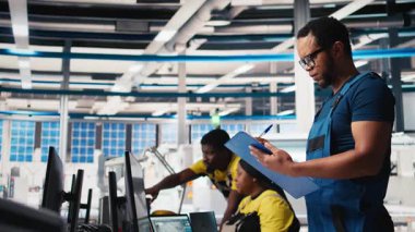 Fotovoltaik fabrikasındaki siyah mühendis güneş panelleri montaj hatlarının performansını inceliyor, ince ayar için evrakları kontrol ediyor. Güneş pili makinelerini denetleyen denetçi. Kamera B.