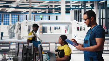 Güneş paneli fabrika teftişinde müfettiş üretim ekipmanları ve üretim süreci, belge dosyalarını kontrol ediyor. Siyahi adam tesiste çevre dostu fotovoltaikleri sağlıyor. Kamera B.