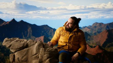 Dağın zirvesine ulaşıp yorgun, dinlenen ve kayalık dağ zirvesinde dinlenen memnun yürüyüşçü. Genç adam nefes kesici manzaranın tadını çıkarıyor. Kamera B.