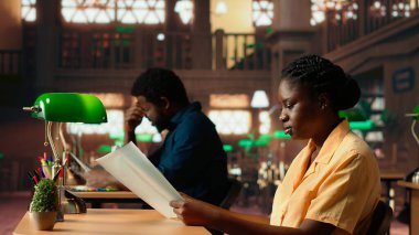 Odaklanmış Afro-Amerikan öğrenci sessiz bir kütüphanede oturup gazete ve ders notları okuyarak yeşil bir lambanın altında ders çalışıyor. Kitaplarla ve öğrenme araçlarıyla çevrili bir kadın. Kamera B.