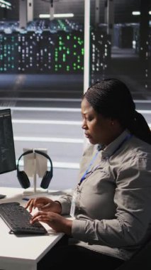Nöro ağ görselleştirmesini izlemek için PC kullanan dikey video veri merkezi sistem yöneticisiName Sunucu odasındaki Afrikalı Amerikalı kadın, bilgisayar kullanarak donanım öğrenme makinelerini denetliyor.