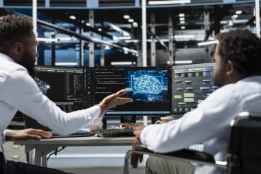 Tekerlekli sandalyedeki adam ve iş arkadaşı yapay zeka ve makine eğitimi için gerekli veri merkezi ekipmanlarını kuruyor. Engelli bilişim uzmanı ve sunucu odasındaki meslektaşı yapay zeka araçları kullanıyor.