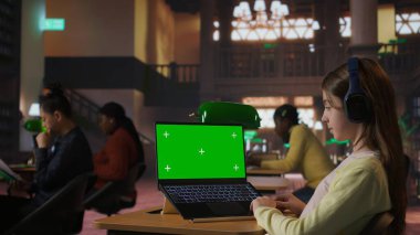 Kütüphanede yeşil ekranlı akademik görevlere odaklanmış liseli kız laptopuyla okul ödevleri üzerinde çalışıyor. Sınıf ödevlerini çözmek, internetten bilgi aramak. Kamera A.