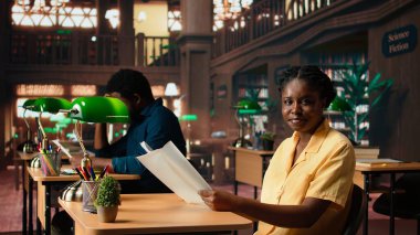 Odaklanmış Afro-Amerikan öğrenci sessiz bir kütüphanede oturup gazete ve ders notları okuyarak yeşil bir lambanın altında ders çalışıyor. Kitaplarla ve öğrenme araçlarıyla çevrili bir kadın. Kamera A.