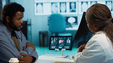 Siyahi erkek hasta, tarama için sıhhiyeye röntgen testi sonuçlarını getiriyor. Teşhis raporlarını inceliyor. Tıbbi uzman kabinedeki radyografi filmini değerlendiriyor. Kamera A.