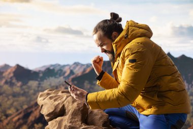 Erkek yürüyüşçü iyi haberler okuyor ve telefonuyla seviniyor. Güçlü sinyal ve internet bağlantısıyla dikkati dağılıyor. Modern kaşif dağ zirvesinde coşkuyla tezahürat yapıyor..
