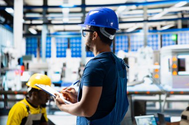 Afrikalı Amerikalı denetçi, dosyalardaki gelişmiş izleme ve üretim araçlarını kullanarak fabrikadaki yenilenebilir enerji ölçümlerini değerlendiriyor. Mühendis güneş enerjisinin doğruluğunu artırıyor.