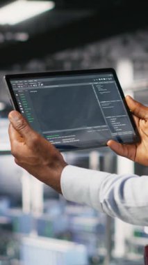Bulanık arkaplandaki yapay zeka görsellemelerini gözden geçiren meslektaşımın yanındaki tablet üzerindeki dikey video veri merkezi kontrol kodu. Bilişim uzmanları sinirsel ağ çıktılarını analiz etmek ve istikrarlı hale getirmek için aygıt kullanırlar