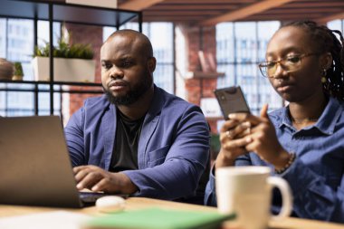 Modern bir dairedeki Afrikalı Amerikalı çift, üretkenliklerini artırmak için teknolojiyi kullanarak bir danışmanlık projesinde işbirliği yapıyor. Adam dizüstü bilgisayarda çalışırken ortağı telefondaki e-postaları cevaplıyor..