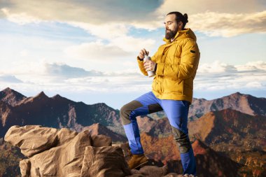 Genç kâşif dağ sırasına tırmanıyor ve su şişesiyle su içiyor. Kayalık dağın tepesinde serinletici bir şeyler içiyor. Yürüyüş aktivitesinden sonra erkek yürüyüşçü ferahlatıcı..