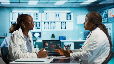 Siyahi pratisyen hekim, sağlık ziyareti sırasında bir hastaya klinik kayıtları ve sonuçları açıklarken, tedavi tavsiyelerine ve tesis ofisinde etkili iletişimlere odaklanıyor. Kamera B.