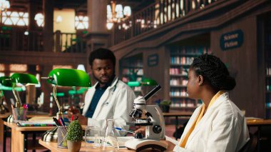 Siyahi öğrenciler mikroskopla virüs ve mikrobiyoloji yöntemlerini araştırıyorlar, kütüphanede fikir ve bilgi alışverişinde bulunuyorlar. Notlar alır ve akademik veri tabanlarına bakar. Kamera A.