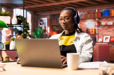 Şirket projesi için verileri araştırırken, kişisel ev işlerinde çalışırken müziği seven bir kişi. Afrika kökenli Amerikalı bir kadın dizüstü bilgisayarın önünde oturmuş kulaklık takıyor.