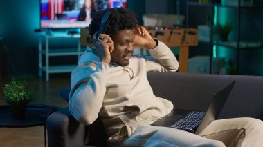 Afrikalı Amerikalı serbest çalışan bir adam internette geziniyor ve müzik dinliyor, modern evindeki şarkılarla eğleniyor. Genç yetişkin kanepede uzanıyor ve oturma odasında dinleniyor. Kamera A.