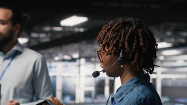 Endüstriyel platformdaki veri merkezi çalışanı mikrofonla konuşurken aşağıdaki yapay zeka platformlarının bakımını denetliyor. Yönetici, derinlemesine öğrenme sistemi görevlerini takım B kamerasıyla koordine etmek için tablet kullanır