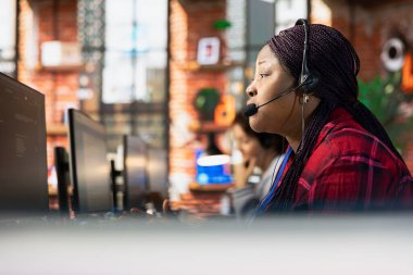 Ofisteki çağrı merkezi temsilcisi soruları cevaplayarak müşterilere yardım ediyor, yakın çekim. Müşteri hizmetlerindeki Afrikalı Amerikalı kadın müşterilerle konuşmak için kulaklık kullanıyor