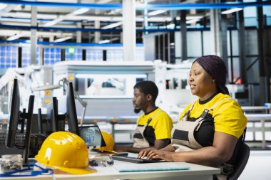 Siyahi kadın mühendis enerji kontrol sistemlerini güneş enerjisi imalatıyla çevrili bir şekilde bilgisayar kurulumunda inceliyor. Kadın uzman fotovoltaik ve endüstriyel üretimi dengeledi.