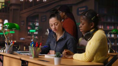 Kütüphane masasında ilkokul kızına rehberlik eden beyaz bir öğretmen, akademik gelişimlerine yardımcı olmak için çalışma kitapları ve notlarla özel dersler veriyor. Öğretmen akademisyenle konuşuyor. Kamera B.