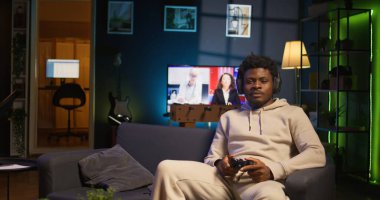 Afrika kökenli yetişkin, takım arkadaşlarıyla konuşmak için kulaklık takıyor ve rahat bir dairede video oyunları oynuyor. Oyuncu oyun konsolunda oyun oynarken internet arkadaşlarıyla sohbet ediyor. Kamera B.