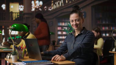 Bilgisayarında akademik kaynaklar ve rehberlik sağlayarak halk kütüphanesinde çalışan kendinden emin bir kütüphanecinin portresi. Veri tabanı içindeki öğrenme materyallerini değerlendiriyorum. Kamera A.