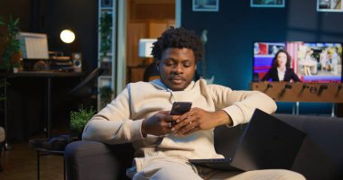 Rahat bir kanepede akıllı telefonuna bakarken gülümseyen rahat bir Afrikalı Amerikalı serbest yazar. Modern ev dekorasyonu şık bir dairede boş zamanların ve çevrimiçi bağlantının altını çizer. Kamera B.
