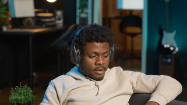 Oturma odasından dizüstü bilgisayara bağlanmış genç bir zenci kanepesinde dinleniyor ve kulaklıkla dinliyor. Neşeli adam günlük işlerini bitirmek için birden fazla iş yapıyor. Kamera A.