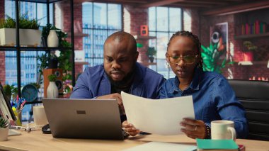 Belgelerindeki takvim randevularını inceleyen iki uzaktan çalışan, programdaki önemli görevleri çözmek için evrakları ve laptop bilgilerini kontrol ediyorlar. Evde işbirliği yapan bir çift. Kamera A.