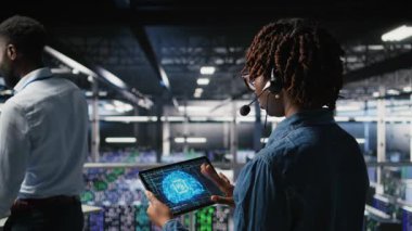 Endüstriyel platformdaki veri merkezi çalışanı mikrofonla konuşurken aşağıdaki yapay zeka platformlarının bakımını denetliyor. Yönetici, derinöğrenme sistemi görevlerini takım, kamera A ile koordine etmek için tablet kullanır