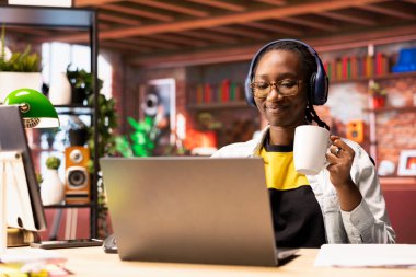 Neşeli bir kadın evde dizüstü bilgisayar başında televizyon seyrediyor ve kahve içiyor. Gülümseyen kişi kulaklık takıyor, apartmanda içecekten keyif alıyor, medya içeriğini tüketiyor