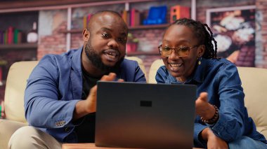 Serbest çalışan çift, yeni bir proje için bilgi toplamak amacıyla evde makaleler okuyarak ve bilgi toplayarak araştırma yapıyor. İnsanlar kanepede birden fazla iş yapıyor ve online raporları inceliyor. Kamera B.