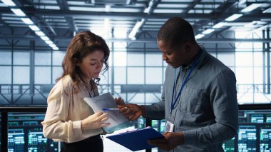 Veri merkezi yöneticileri evrak dosyalarını okuyor, sistem tanılarını gözden geçiriyorlar. Sunucu odası çalışanları bakım programı belgelerini ve siber güvenlik protokollerini inceliyor.