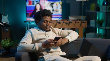 Rahat bir kanepede akıllı telefonuna bakarken gülümseyen rahat bir Afrikalı Amerikalı serbest yazar. Modern ev dekorasyonu şık bir dairede boş zamanların ve çevrimiçi bağlantının altını çizer. Kamera A.