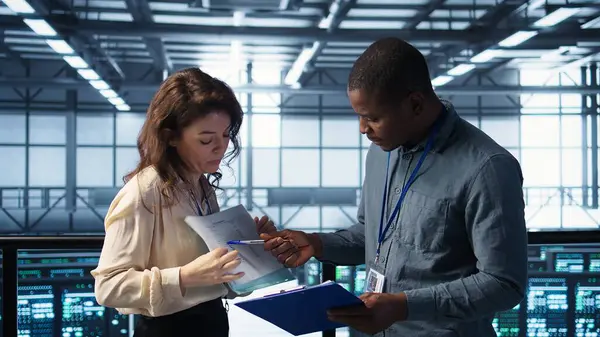 Veri merkezi yöneticileri evrak dosyalarını okuyor, sistem tanılarını gözden geçiriyorlar. Sunucu odası çalışanları bakım programı belgelerini ve siber güvenlik protokollerini inceliyor.