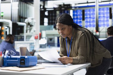 Kadın mühendis kağıtlardaki üretim hattı verilerini inceliyor, yenilenebilir enerji teknolojisine etkili kalite kontrolü ve bağlılığı sağlamak için güneş paneli fabrikasında fabrika faaliyetlerini yönetiyor..