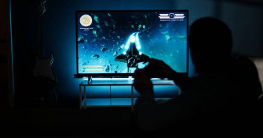 Genç yetişkin, akıllı televizyonunda video oyunlarıyla eğleniyor. Oturma odasındaki LCD monitörüne konsolunu bağlıyor ve evde oyun gecesinin tadını çıkarıyor. Afrikalı Amerikalı oyuncu. Kamera A.