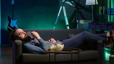 Gülümseyen adam evde kanepede uzanmış patlamış mısır yerken televizyon kanalında yayınlanan filmlere göz gezdiriyordu, işten izin almanın keyfini çıkarıyordu. Apartman dairesine uzanmış abur cubur yiyen adam, kamera A.